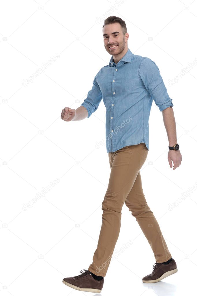 Side view of handsome casual man smiling while wearing blue shirt, stepping on white studio background