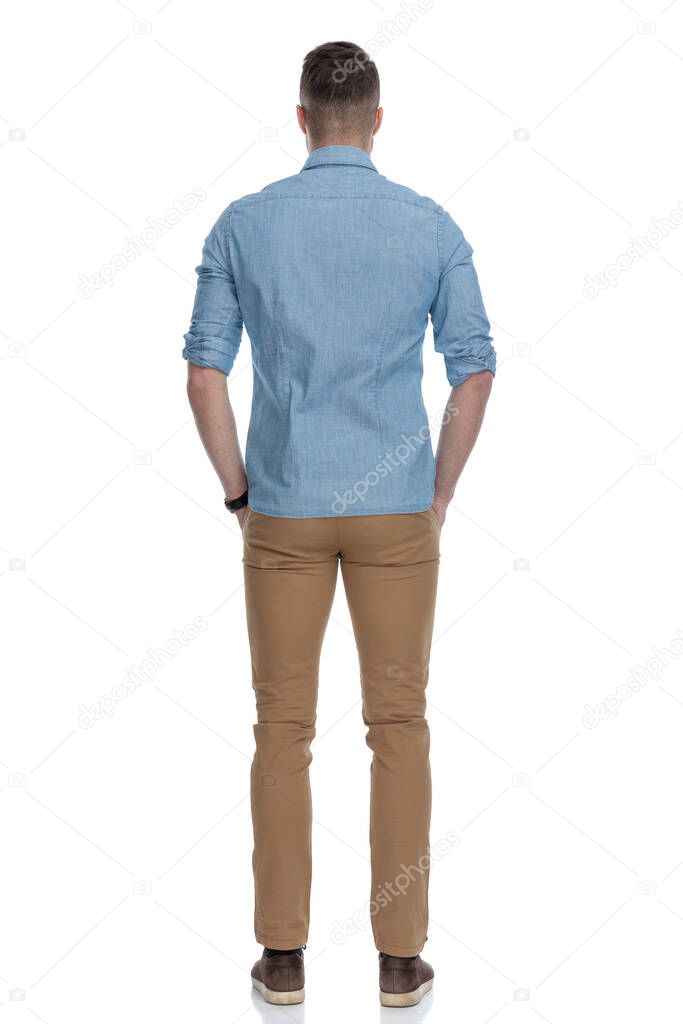 Rear view of casual man holding his hands in his pockets while wearing blue shirt, standing on white studio background