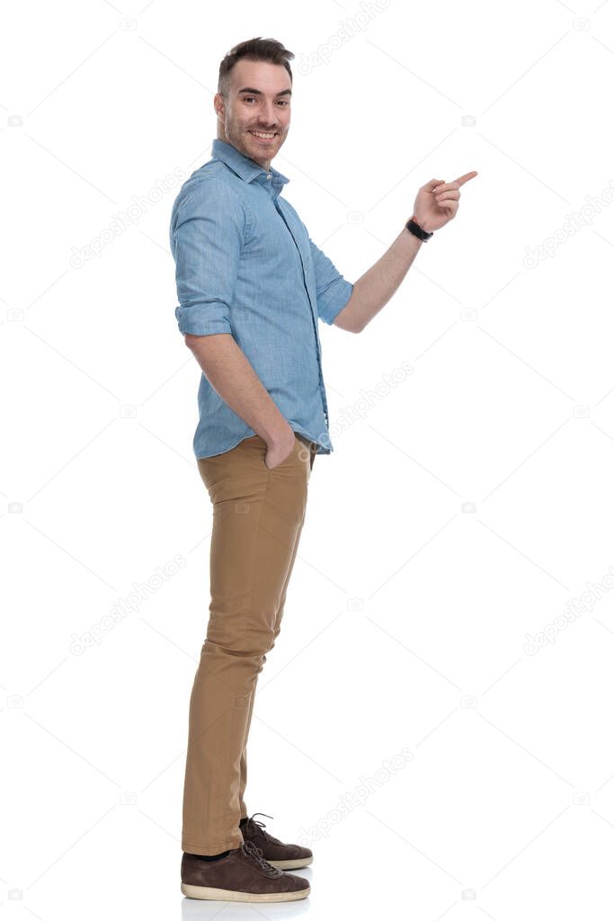 Side view of happy casual man pointing with hand in pocket while wearing blue shirt, standing on white studio background
