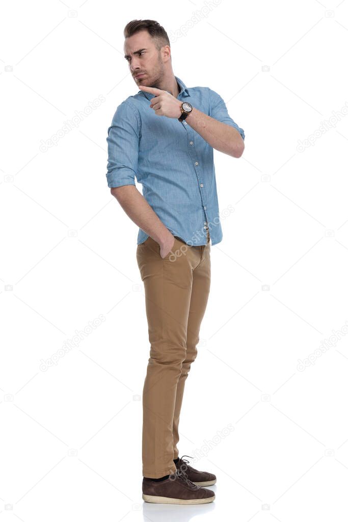 Side view of upset casual man pointing and frowning while wearing blue shirt, standing on white studio background