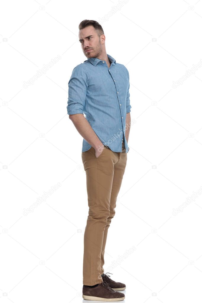 Side view of bothered casual man frowning and looking over shoulder while wearing blue shirt, standing on white studio background
