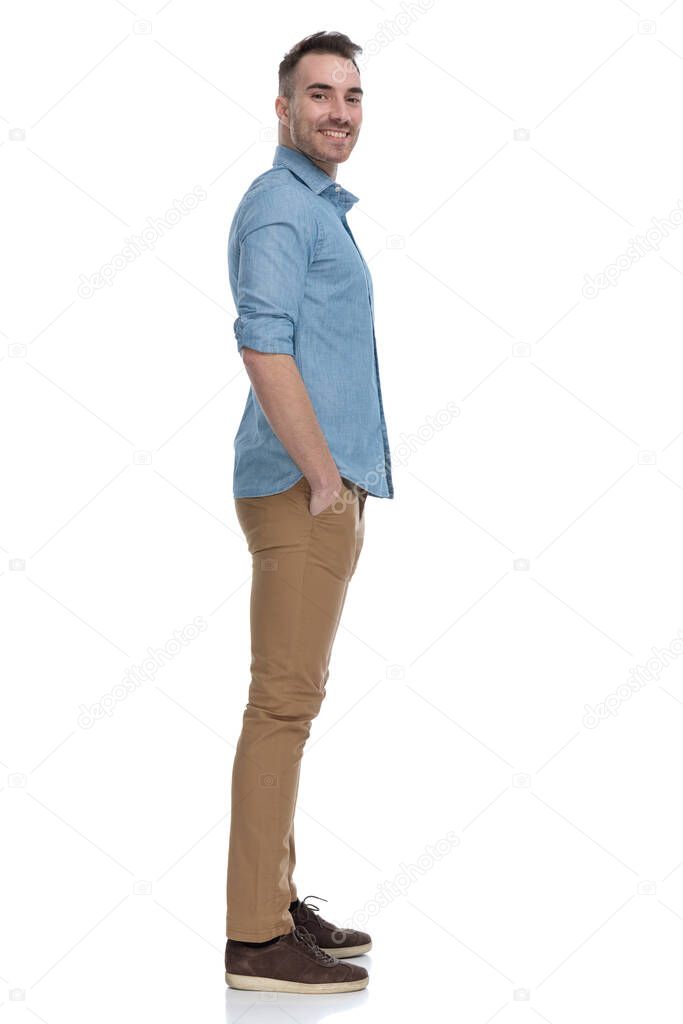 Side view of happy  casual man smiling with both hands in pockets while wearing blue shirt, standing on white studio background