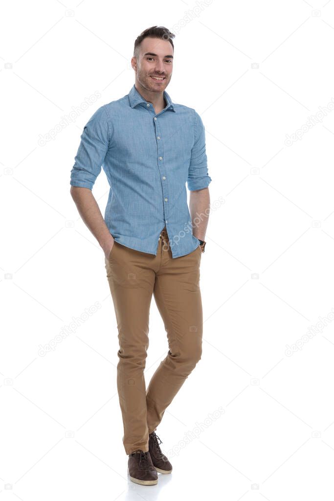Positive casual man smiling with both hand in his pockets while wearing blue shirt, standing on white studio background