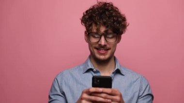 Gözlüklü, gözlüklü ve telefonuna mesaj atan yakışıklı bir adam pembe stüdyo arka planında mutlu bir şekilde başparmak kaldırıyor.
