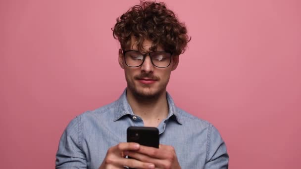 若いカジュアルな男が眼鏡をかけて電話で読み ピンクのスタジオの背景に幸せを指して — ストック動画