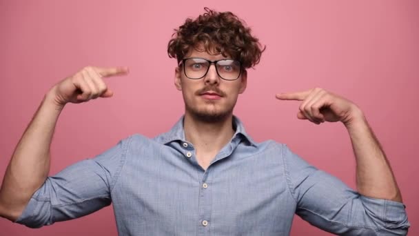 眼鏡をかけた魅力的なカジュアルな男が立ち上がり狂気のジェスチャーをしてからピンクのスタジオの背景で笑う — ストック動画