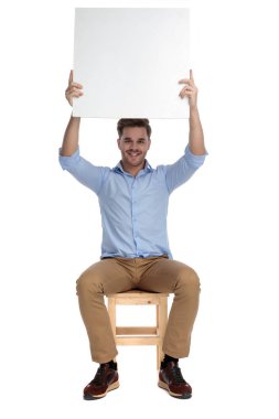 Beyaz stüdyo arka planında bir sandalyede otururken elinde reklam panosu tutan ve gülümseyen mutlu bir adam.