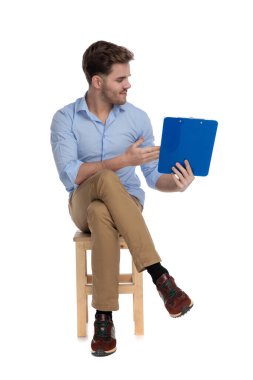 Beyaz stüdyo arka planında bir sandalyede otururken pano sunan sıradan bir genç.