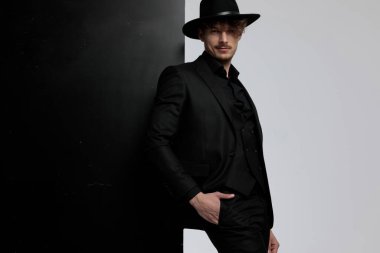 Charming fashion model posing with his hand in his pocket while wearing suit and hat, standing on black and white studio background clipart