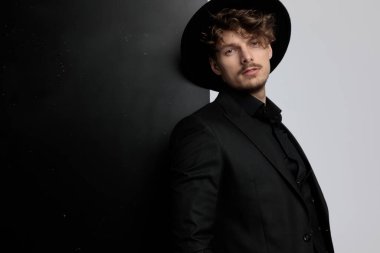 Çekici manken takım elbise ve şapka giyerken ileri bakıyor, siyah beyaz stüdyo arka planında duruyor.