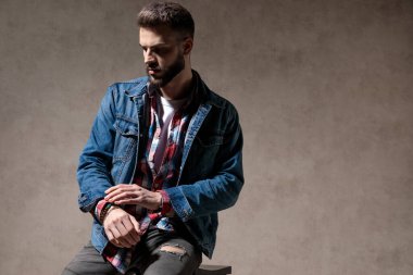 Düşünceli bir model kot ceket giyer ve stüdyo arka planında bir taburede otururken aşağı bakar ve düşünür.