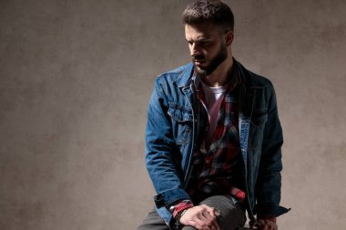 Rahatsız bir manken kot ceket giyerken ve arka planda duvar kâğıdında bir taburede otururken aşağılara bakıyor ve kaşlarını çatıyor.