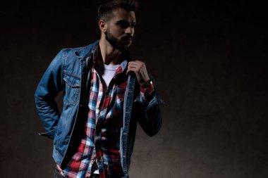 Esrarengiz bir model, kot ceket giyerken ve arka planda duvar kâğıdı üzerinde dururken yakasını arkadan düzeltiyor.