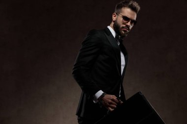 Sıkıntılı moda işadamı takım elbise ve güneş gözlüğü takarken evrak çantası tutuyor, duvar kağıdı stüdyosunda yürüyor.