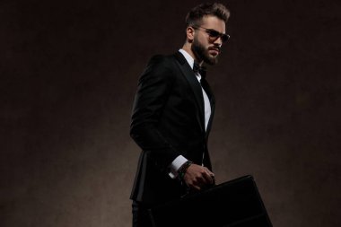 Moda işadamı takım elbise ve güneş gözlüğü takarken evrak çantası tutuyor, duvar kağıdı stüdyosunda yürüyor.