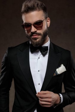 Takım elbise ve güneş gözlüğü takarken gülümseyen, duvar kağıdı stüdyosunun arka planında duran pozitif moda damadı.