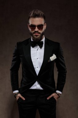 Yakışıklı damat, elleri ceplerinde, takım elbise ve güneş gözlüğü takmış, duvar kâğıdı stüdyo arka planında duruyor.