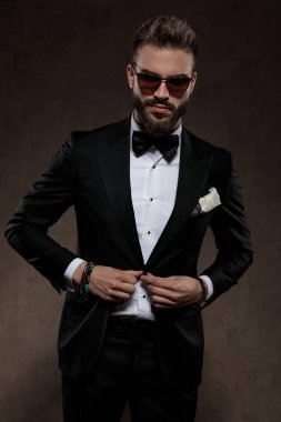 Takım elbise ve güneş gözlüğü takıp, arka planda duran ceketinin düğmelerini açıp gülümseyen moda damadı.