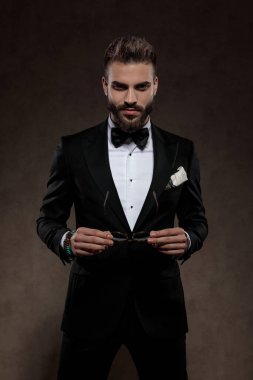 Ciddi moda damadı takım elbise giyerken güneş gözlüğünü çıkarıyor, duvar kağıdı stüdyosunun arka planında duruyor.