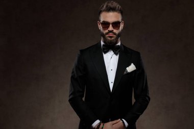 Ciddi moda damadı güneş gözlüğü ve takım elbise takıp, arka planda duvar kâğıdı stüdyosunda dikilirken ileriye bakıyor.