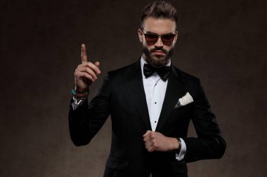 Kendine güvenen moda damadı, güneş gözlüğü ve takım elbise takıp, arka planda duvar kâğıdının üstünde dikiliyor.