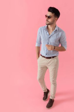 Gömlek ve güneş gözlüğü takmış, pembe stüdyo arka planında yürürken cebinde el ele bakan hevesli, akıllı, sıradan bir adam.