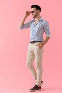 Kendine güveni tam, rahat bir adam gömlek giyerken cebinde güneş gözlüğünü çıkarıyor, pembe stüdyo arka planında yürüyor.