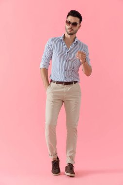 Tişört ve güneş gözlüğü takıp, pembe stüdyo arka planında yürürken cebinde duran havalı, akıllı, sıradan bir adam.