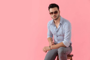 Güneş gözlüğü ve gömlek takmış, pembe stüdyo arka planında bir taburede otururken ileriye bakan sert, akıllı, sıradan bir adam.