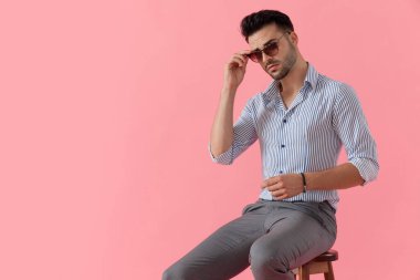 Yakışıklı, sıradan bir adam, gömlek giyerken güneş gözlüğünü ayarlıyor, pembe stüdyo arka planında bir taburede oturuyor.