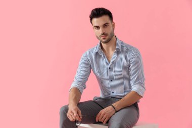 Çekici, zeki, sıradan bir adam gözlüğünü gömlek giyerken, pembe stüdyo arka planında bir taburede otururken tutuyor.