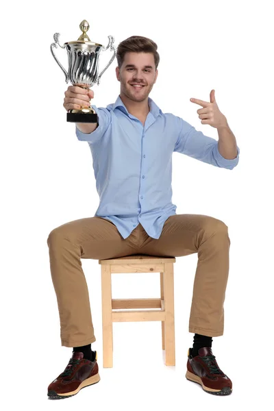 快乐而随意的男人拿着奖杯 一边指着奖杯 一边坐在一张背景为白色的椅子上 — 图库照片