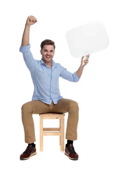 陽気なカジュアルな男を保持スピーチバブルとお祝いながら 白いスタジオの背景に椅子に座って — ストック写真