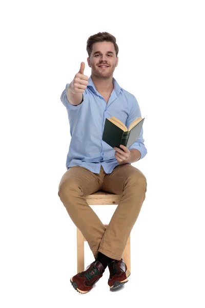 积极而随意的男人一边看书 一边竖起大拇指 一边笑着坐在白色画室的椅子上 — 图库照片