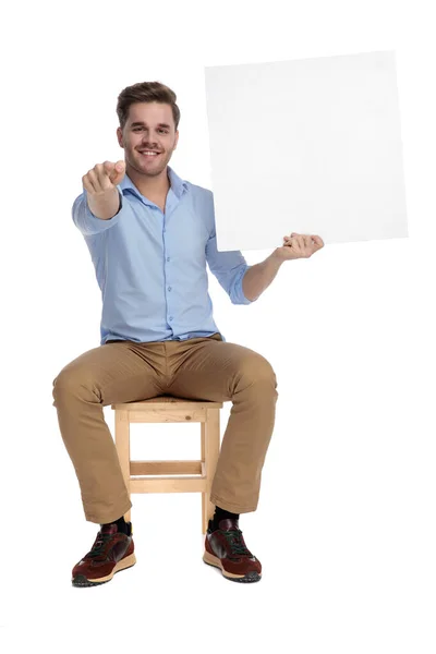 白いスタジオの背景に椅子に座っている間に笑顔で 壁紙を保持し 前方を指して幸せなカジュアルな男 — ストック写真