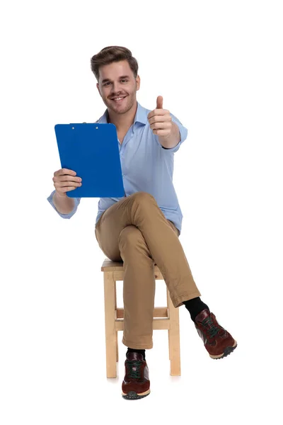 快乐而随意的男人拿着剪贴板 摆出一副不错的姿势 坐在白色画室背景的椅子上笑着 — 图库照片