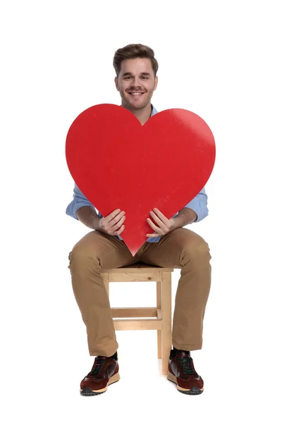 幸せカジュアルな男を保持心の形と笑顔ながら 上の椅子に座っています白いスタジオの背景 — ストック写真