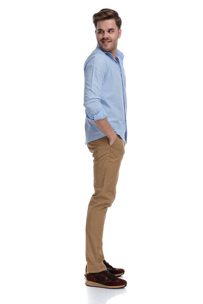 Вид сбоку на счастливого случайного человека, оглядывающегося через плечо с руками в карманах в рубашке и стоящего на белом фоне студии
