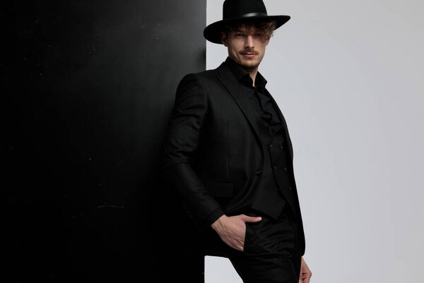 Charming fashion model posing with his hand in his pocket while wearing suit and hat, standing on black and white studio background