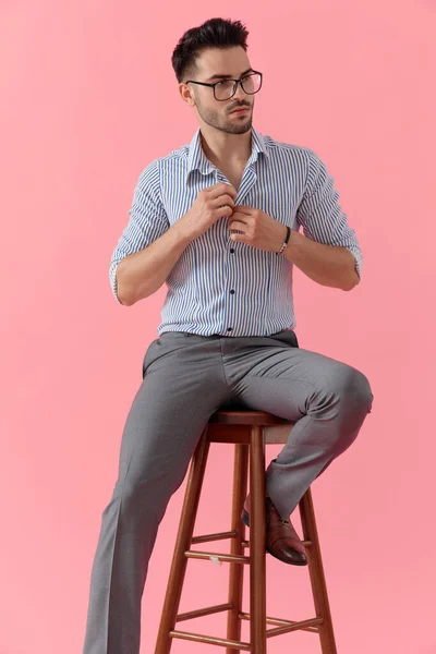 眼鏡をかけながらシャツのボタンを外しピンクのスタジオの背景にあるスツールに座っています — ストック写真