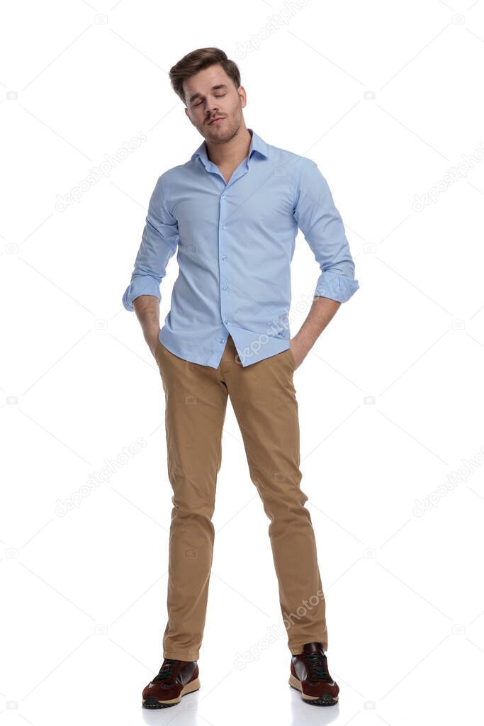 Tired casual man holding eyes closed and hands in pockets while wearing shirt and standing on white studio background