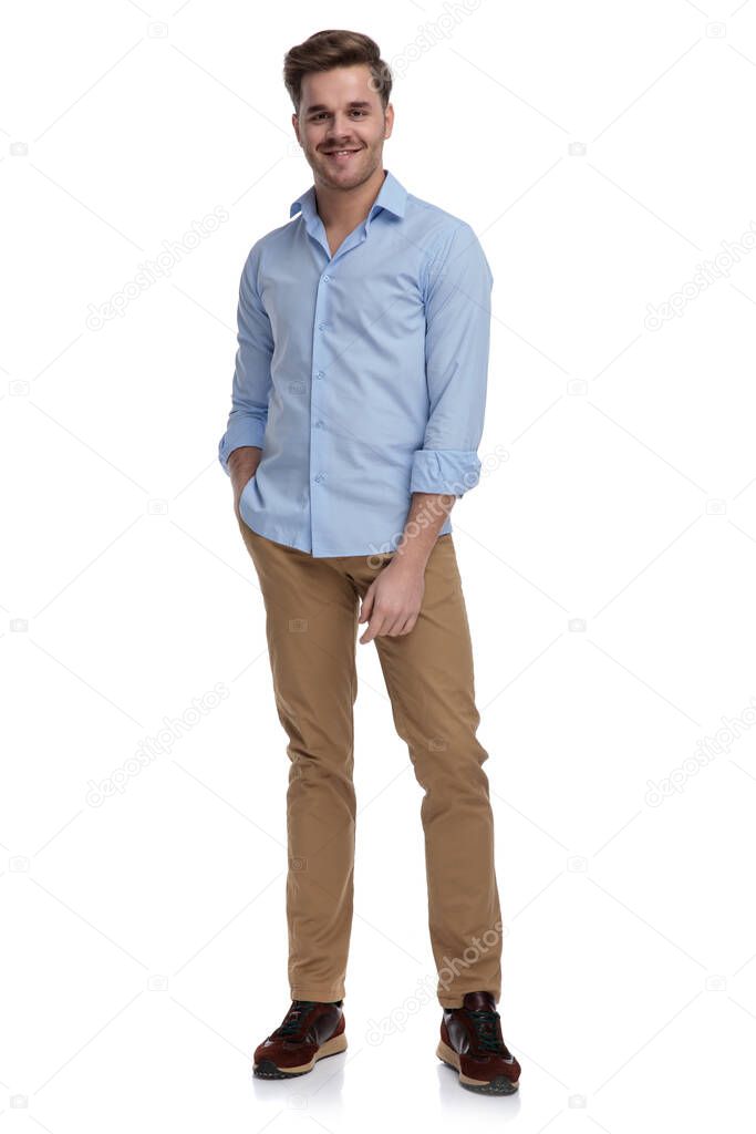 Positive casual man smiling with hand in pocket while wearing shirt and standing on white studio background