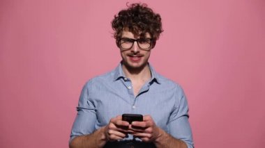 Gözlüklü ve telefonla mesaj atan genç bir adam pembe stüdyo arka planında mutlu bir şekilde başparmak kaldırıyor.