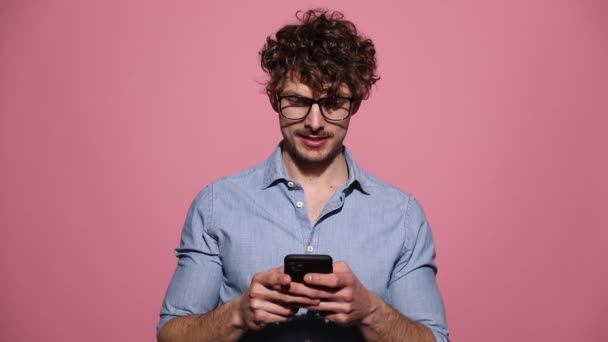 眼鏡をかけ電話でテキストメッセージを送る魅力的なカジュアルな男が驚いてピンクのスタジオの背景で成功を祝う — ストック動画