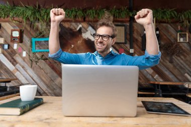 Gözlüklü sıradan bir adam masada oturuyor ve kahvecide heyecanlı bir şekilde dizüstü bilgisayarının önünde başarısını kutluyor.