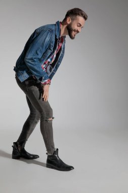 Kot ceket giyen yakışıklı bir adamın yan görünüşü gri stüdyo arka planında eğilmiş ve gülerken mutlu.