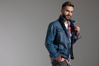 Ekose gömlekli genç bir adamın yan görünüşü. Bir eli cebinde duruyor ve ceketini düzeltiyor. Diğeri gri stüdyo arka planında.