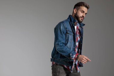 Gri stüdyo arka planında ciddi bir tavırla poz verirken kot ceket giyen ve parmağıyla işaret eden yakışıklı bir adamın yan görünüşü.