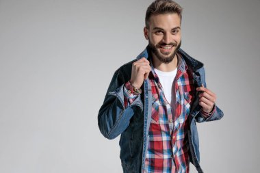 Gri stüdyo arka planında mutlu bir şekilde gülümserken ekose gömlek giyen ve kot ceketini düzelten çekici bir adam.