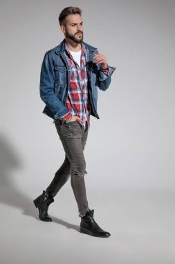 Gri stüdyo arka planında, tek eli cebinde yürüyen sıradan, çekici bir adamın yan görünüşü.